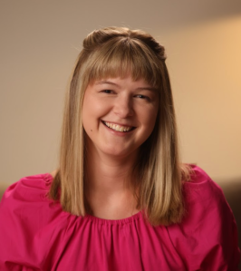 A woman with straight blonde hair, wearing a pink blouse, smiles at the camera.